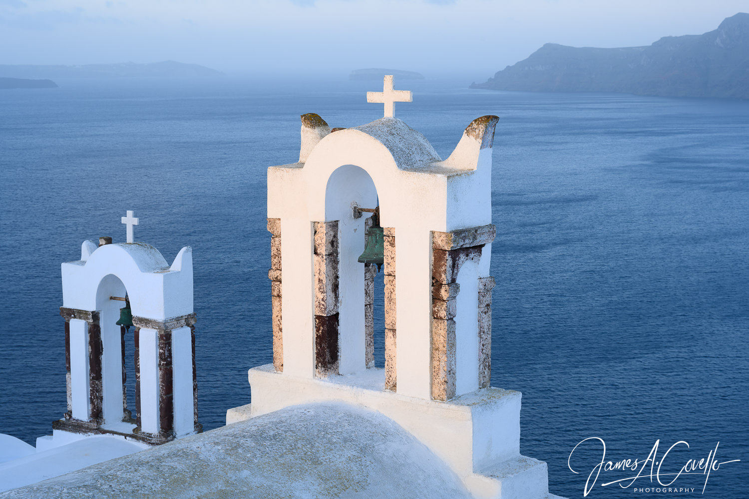 The sea sits behind two white belltowers with crosses on their tops.
